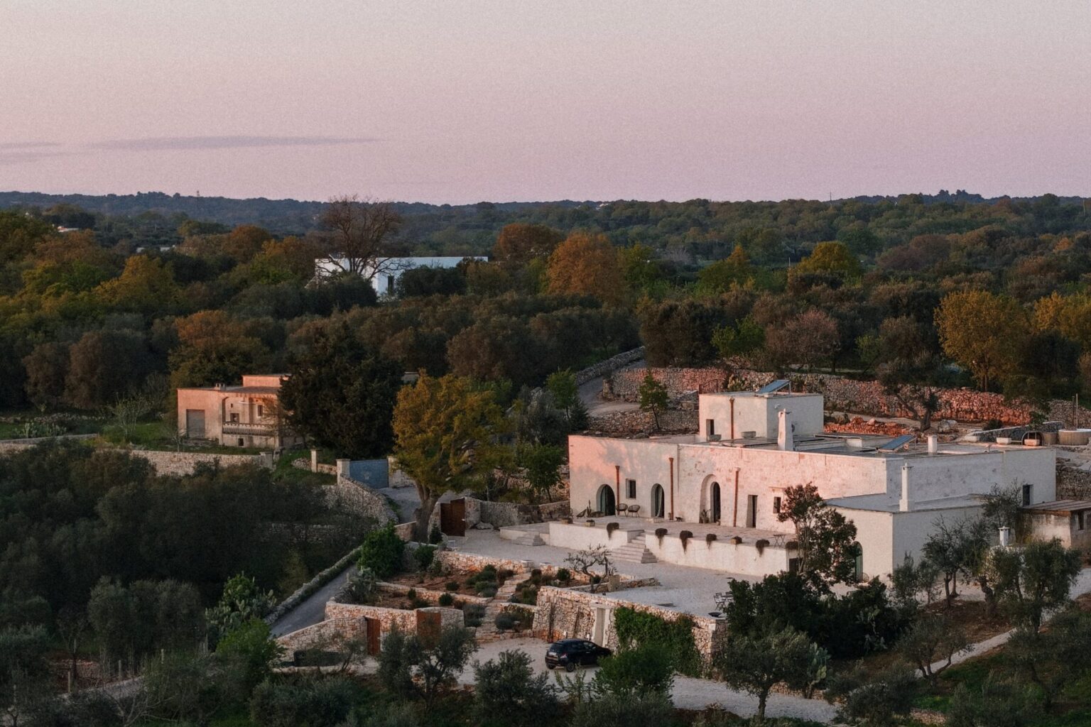 Aerial view of a modern white villa surrounded by lush greenery and olive trees, set against a serene sunset sky. The property features landscaped gardens, a stone pathway, and outdoor seating areas, showcasing a harmonious blend of nature and contemporary architecture. | Only on SopranoVillas