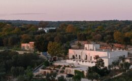 Aerial view of a modern white villa surrounded by lush greenery and olive trees, set against a serene sunset sky. The property features landscaped gardens, a stone pathway, and outdoor seating areas, showcasing a harmonious blend of nature and contemporary architecture.
