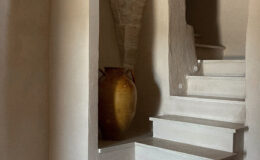 Interior staircase leading to an upper level, featuring whitewashed walls and stone archways. A decorative ceramic vase sits at the base of the stairs, adding a rustic touch to the serene ambiance of the space. Natural light highlights the textures of the walls and flooring.