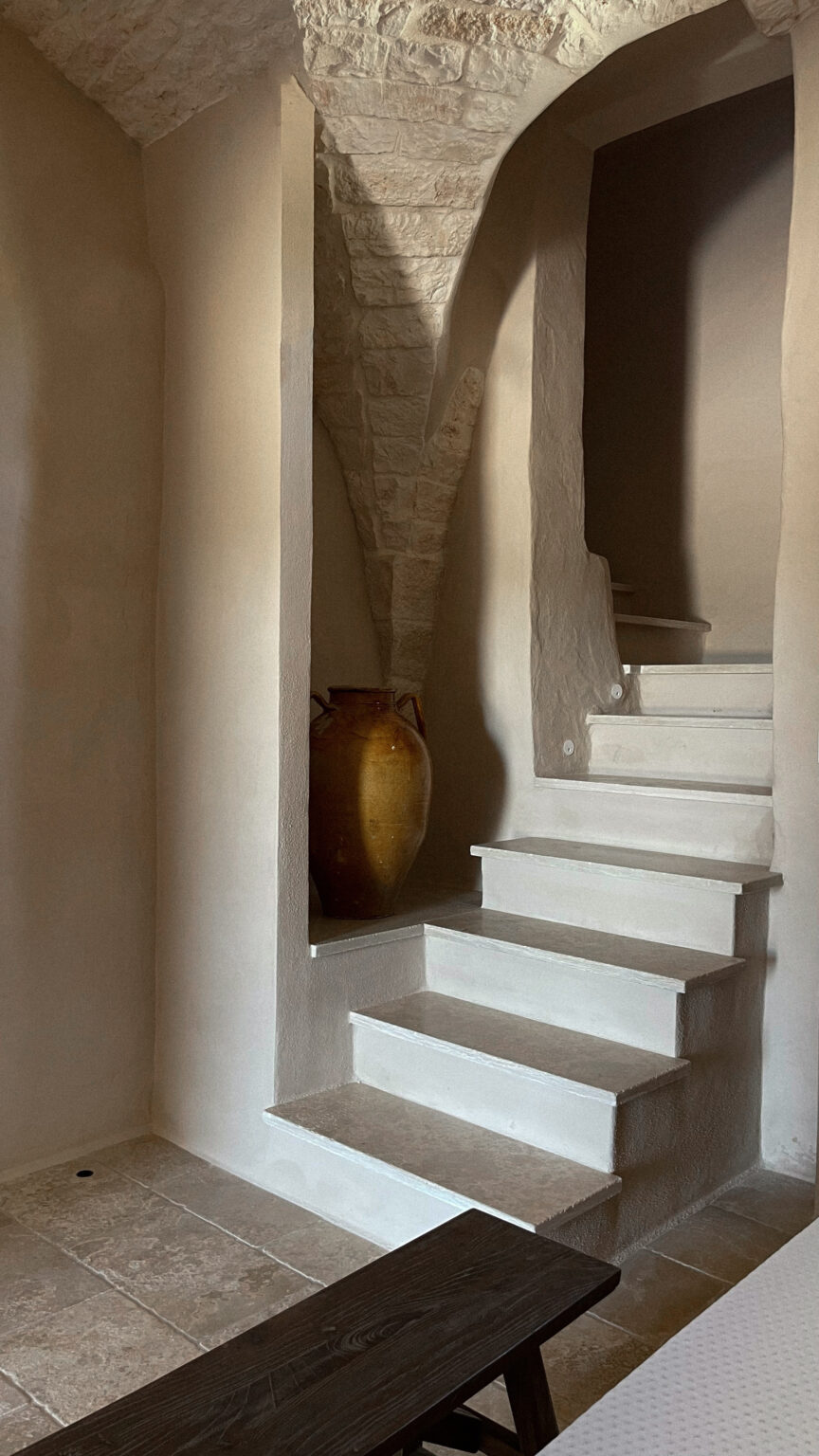 Interior staircase leading to an upper level, featuring whitewashed walls and stone archways. A decorative ceramic vase sits at the base of the stairs, adding a rustic touch to the serene ambiance of the space. Natural light highlights the textures of the walls and flooring. | Only on SopranoVillas