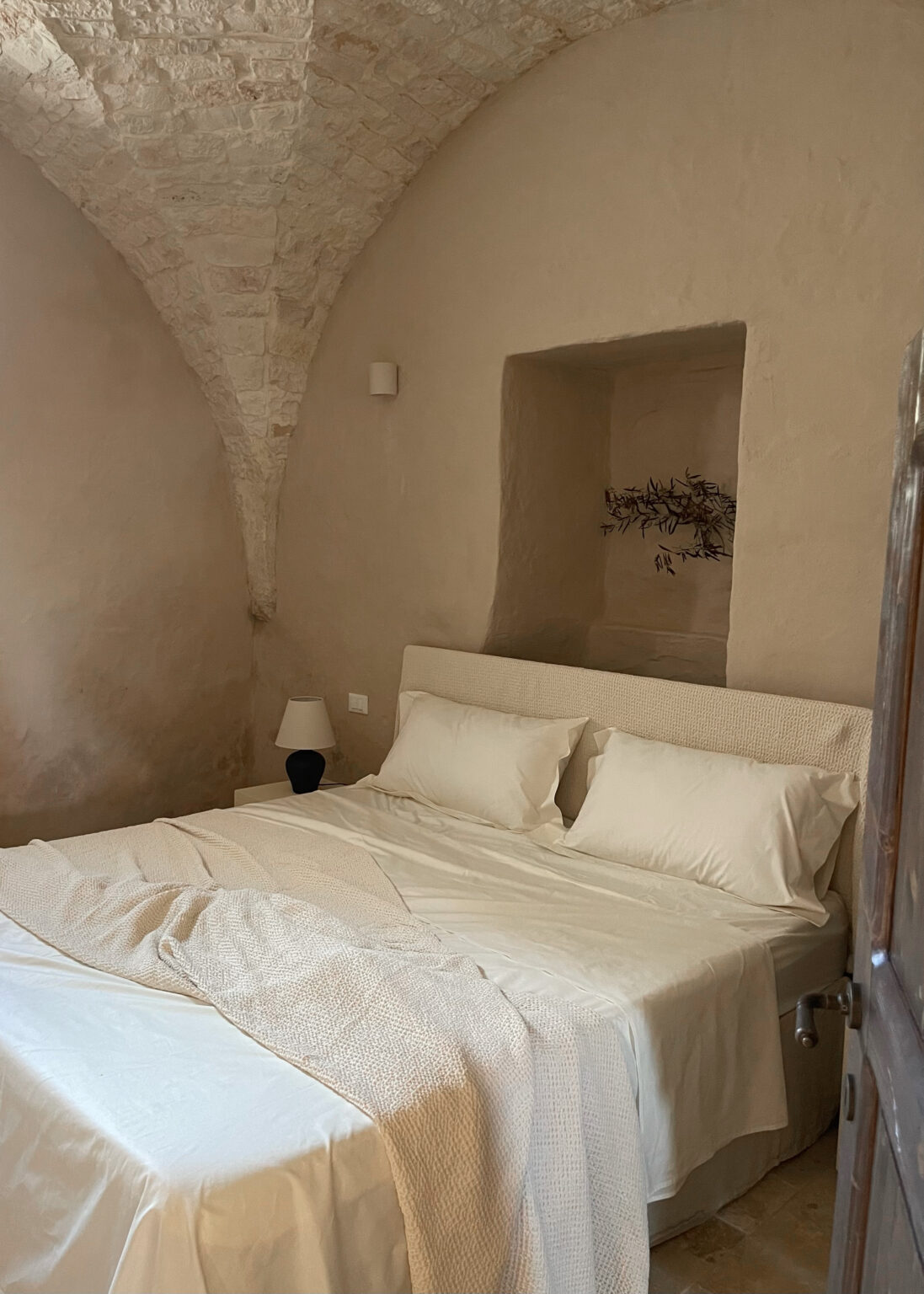 Cozy bedroom featuring a minimalist design with an arched stone ceiling, a plush bed with white linens, and a soft throw blanket. The room includes a bedside lamp and a decorative wall niche with a natural element, creating a serene and inviting atmosphere. Ideal for relaxation and comfort. | Only on SopranoVillas
