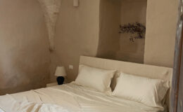 Cozy bedroom featuring a minimalist design with an arched stone ceiling, a plush bed with white linens, and a soft throw blanket. The room includes a bedside lamp and a decorative wall niche with a natural element, creating a serene and inviting atmosphere. Ideal for relaxation and comfort.