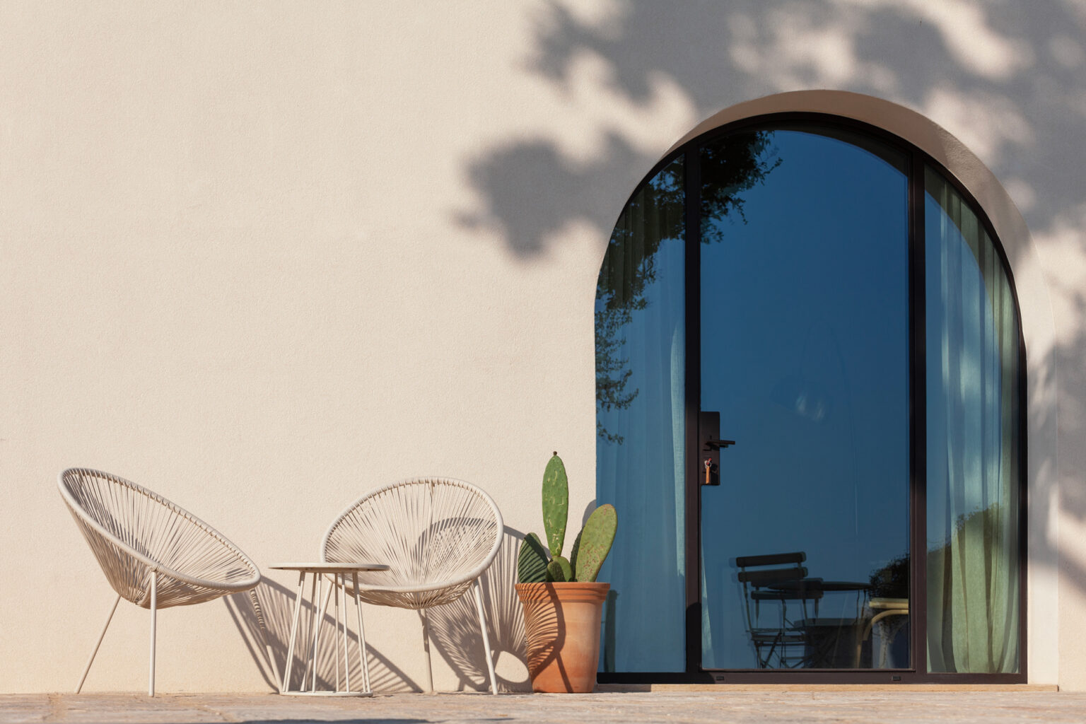 Stylish outdoor seating area featuring two woven chairs and a small table next to a potted cactus, with a large arched glass door reflecting sunlight and greenery. Perfect for modern home design and outdoor relaxation. | Only on SopranoVillas