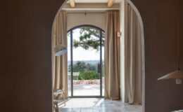 Cozy bedroom interior featuring a textured linen bedspread, with an arched doorway leading to a serene outdoor view. Natural light filters through sheer curtains, illuminating the space and highlighting wooden beams and minimalist decor.