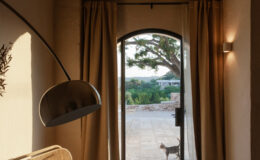 Cozy interior featuring a modern chair and stylish floor lamp, with a large arched window showcasing a view of a stone patio and greenery outside, while a cat walks by. Natural light illuminates the space, highlighting the warm tones of the decor and furnishings.