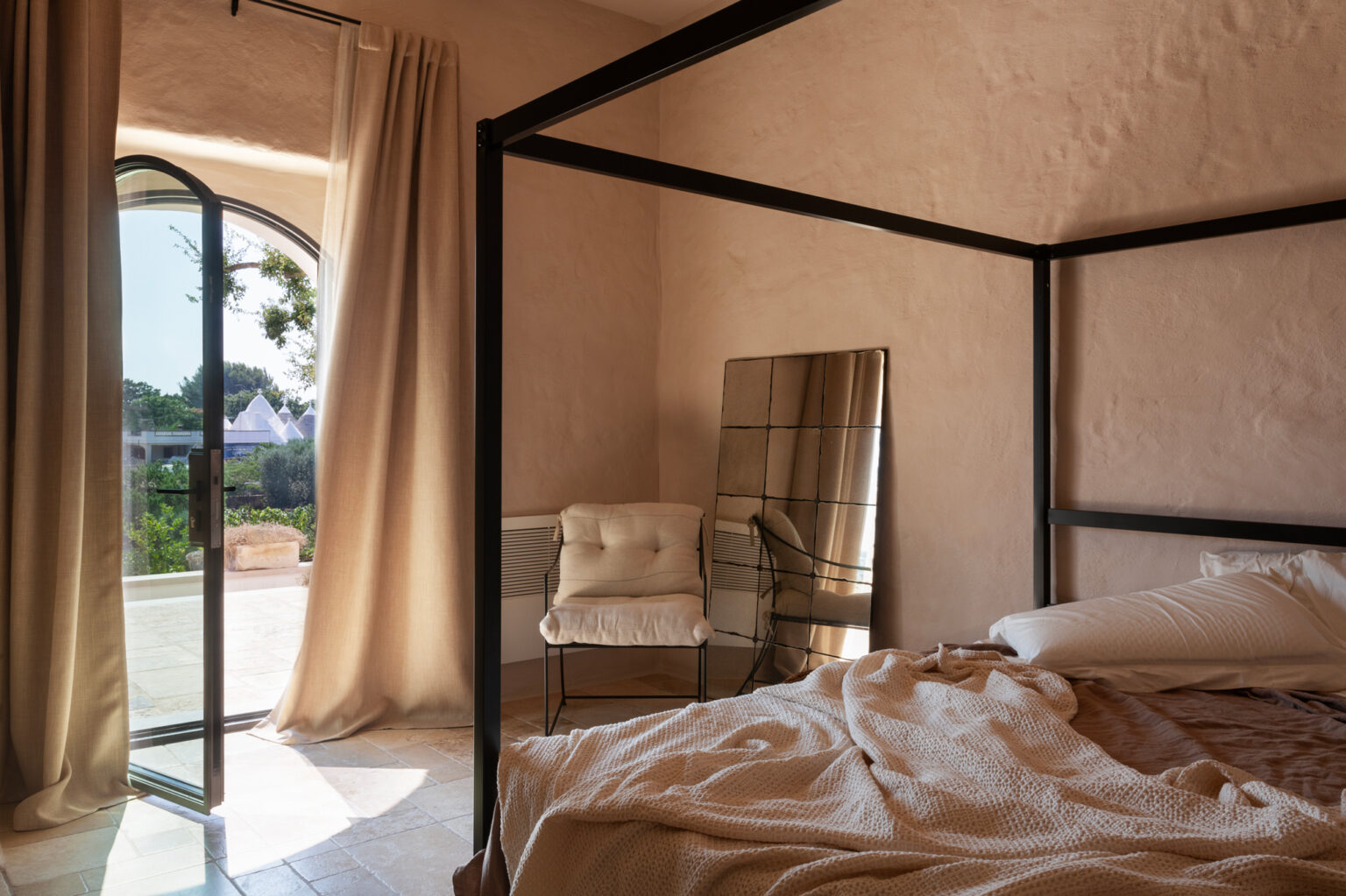 Modern bedroom interior featuring a four-poster bed, a cozy chair, and a large mirror. Soft natural light filters through an arched window, illuminating the textured walls and neutral color palette, creating a serene and inviting atmosphere. | Only on SopranoVillas
