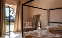 Modern bedroom interior featuring a four-poster bed, a cozy chair, and a large mirror. Soft natural light filters through an arched window, illuminating the textured walls and neutral color palette, creating a serene and inviting atmosphere.