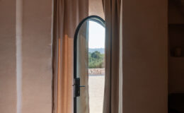 Interior view of a minimalist bedroom featuring a modern arched door opening to an outdoor landscape, with soft curtains framing the entrance, stone flooring, and a cozy bed setup.