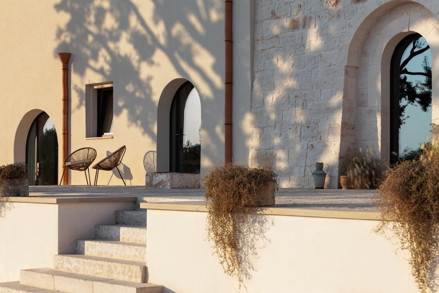 Modern architectural exterior featuring a textured stone wall, arched windows, and stylish outdoor seating. The scene includes two woven chairs on a spacious terrace, surrounded by potted plants and soft evening sunlight casting shadows. Ideal for showcasing contemporary outdoor living spaces. | Only on SopranoVillas