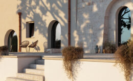 Modern architectural exterior featuring a textured stone wall, arched windows, and stylish outdoor seating. The scene includes two woven chairs on a spacious terrace, surrounded by potted plants and soft evening sunlight casting shadows. Ideal for showcasing contemporary outdoor living spaces.
