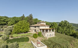 Aerial view of a charming stone villa surrounded by lush greenery and trees, featuring a spacious patio and well-maintained garden, set against a clear blue sky. Ideal for a tranquil retreat in nature.