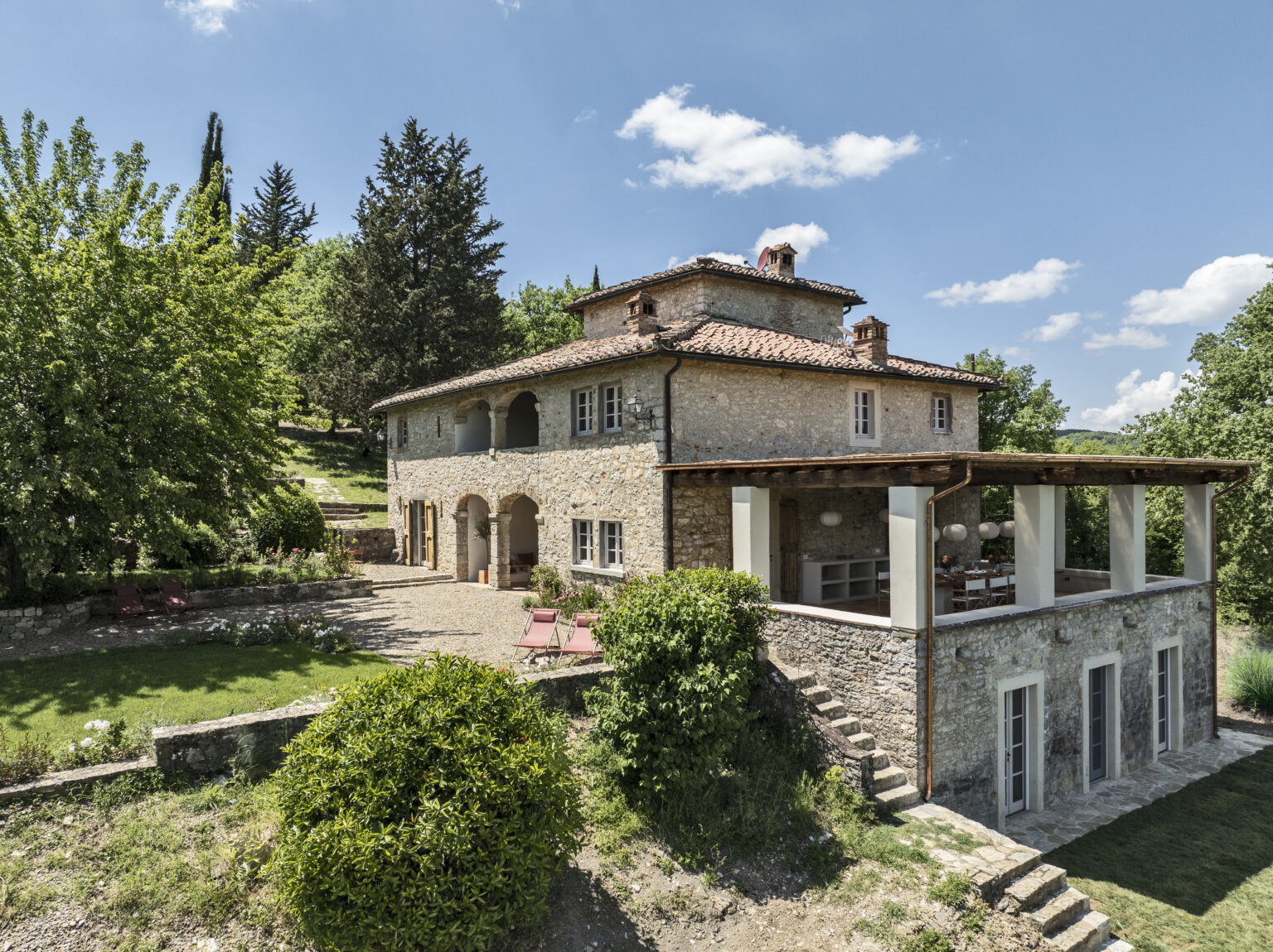 Charming stone villa surrounded by lush greenery, featuring a spacious outdoor area with seating and a picturesque view, ideal for relaxing or entertaining. The property showcases traditional architecture with modern elements, set against a clear blue sky. | Only on SopranoVillas