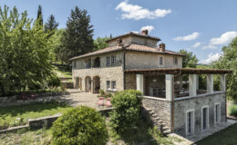 Charming stone villa surrounded by lush greenery, featuring a spacious outdoor area with seating and a picturesque view, ideal for relaxing or entertaining. The property showcases traditional architecture with modern elements, set against a clear blue sky.