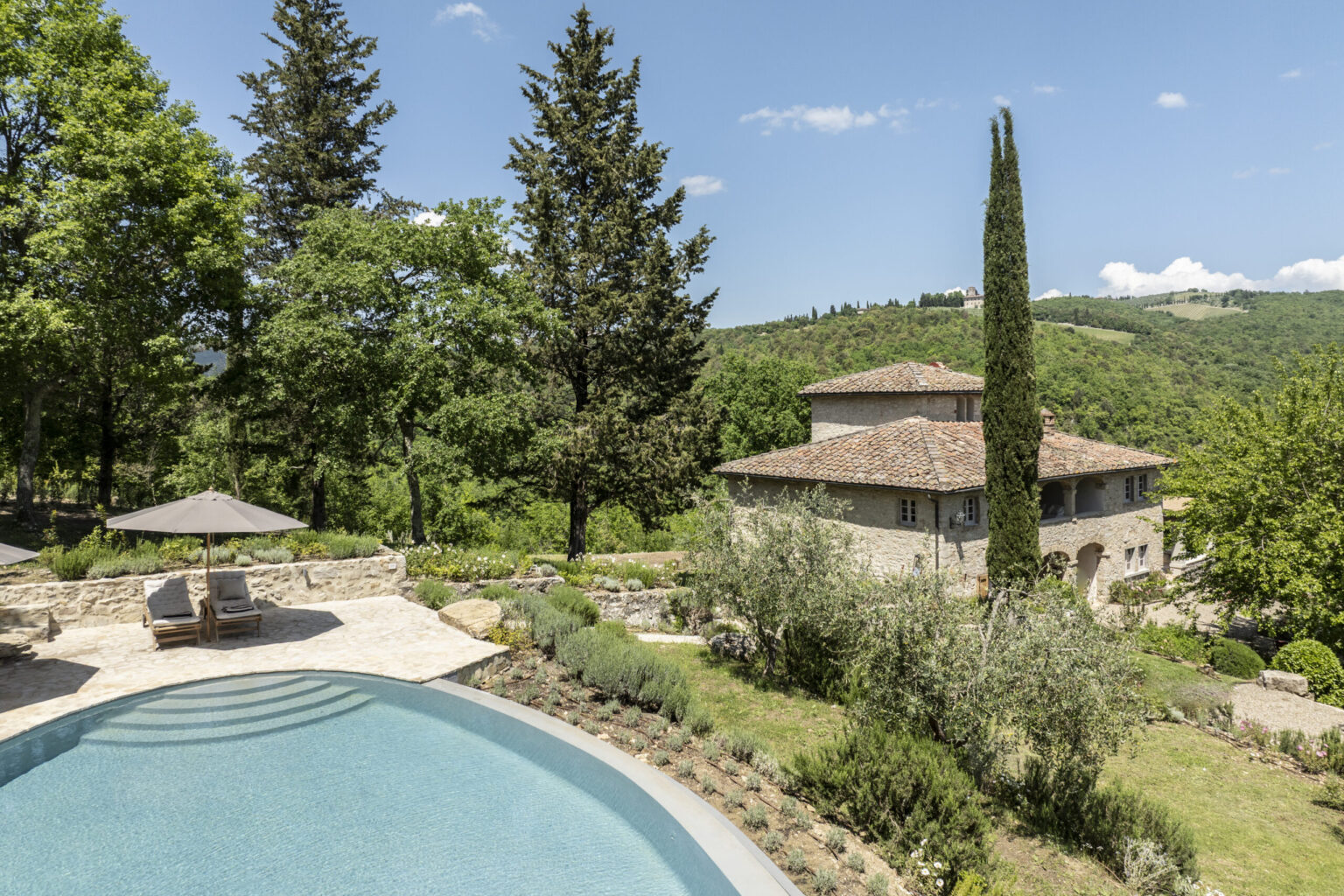 Scenic view of a luxurious villa with a swimming pool surrounded by lush greenery and trees, set against a backdrop of rolling hills under a clear blue sky. The property features a rustic stone exterior, comfortable lounge chairs under umbrellas, and landscaped gardens. Ideal for relaxation and enjoying nature. | Only on SopranoVillas