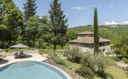 Scenic view of a luxurious villa with a swimming pool surrounded by lush greenery and trees, set against a backdrop of rolling hills under a clear blue sky. The property features a rustic stone exterior, comfortable lounge chairs under umbrellas, and landscaped gardens. Ideal for relaxation and enjoying nature.