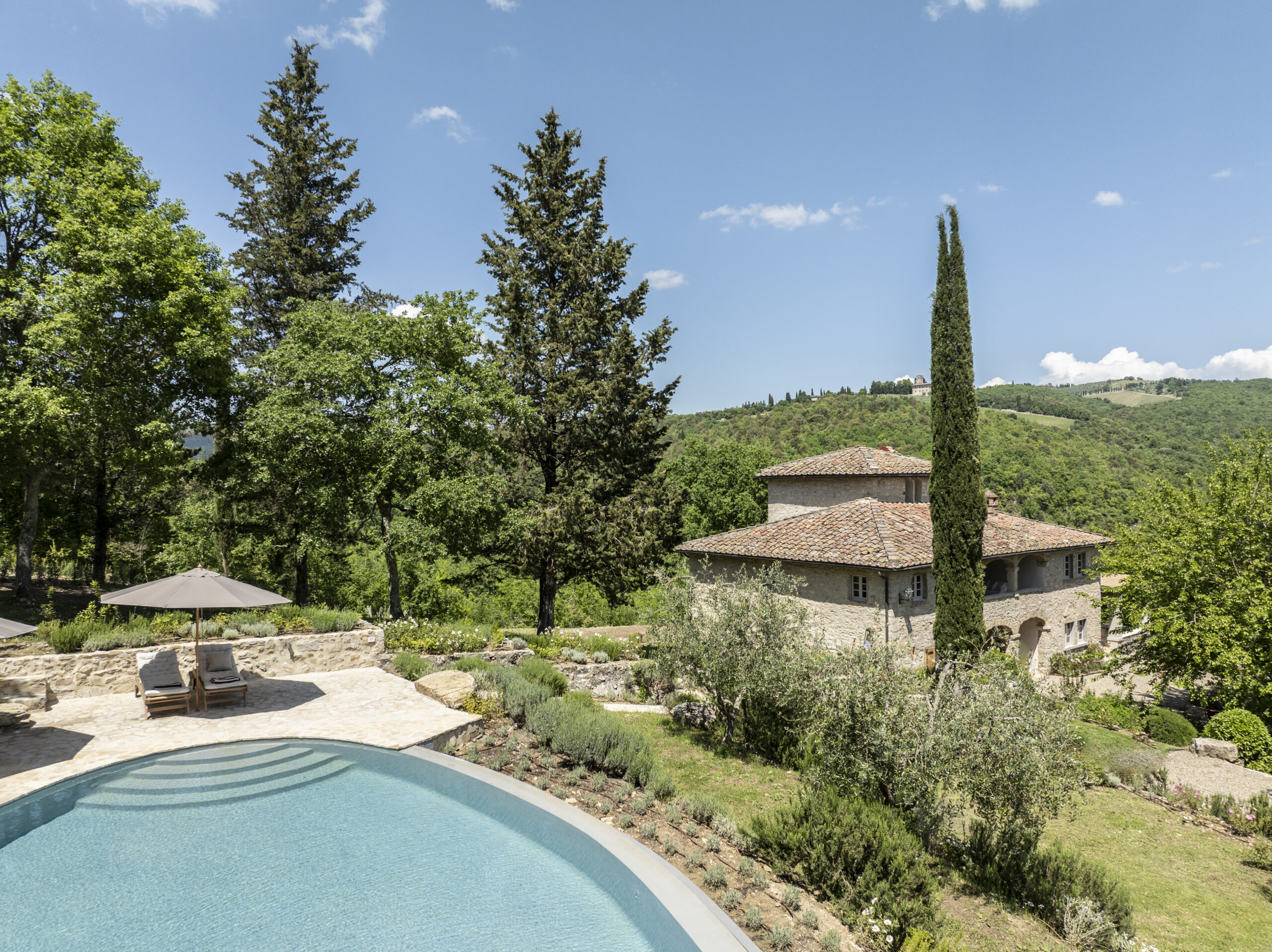 Scenic view of a luxurious villa with a swimming pool surrounded by lush greenery and trees, set against a backdrop of rolling hills under a clear blue sky. The property features a rustic stone exterior, comfortable lounge chairs under umbrellas, and landscaped gardens. Ideal for relaxation and enjoying nature. | Only on SopranoVillas