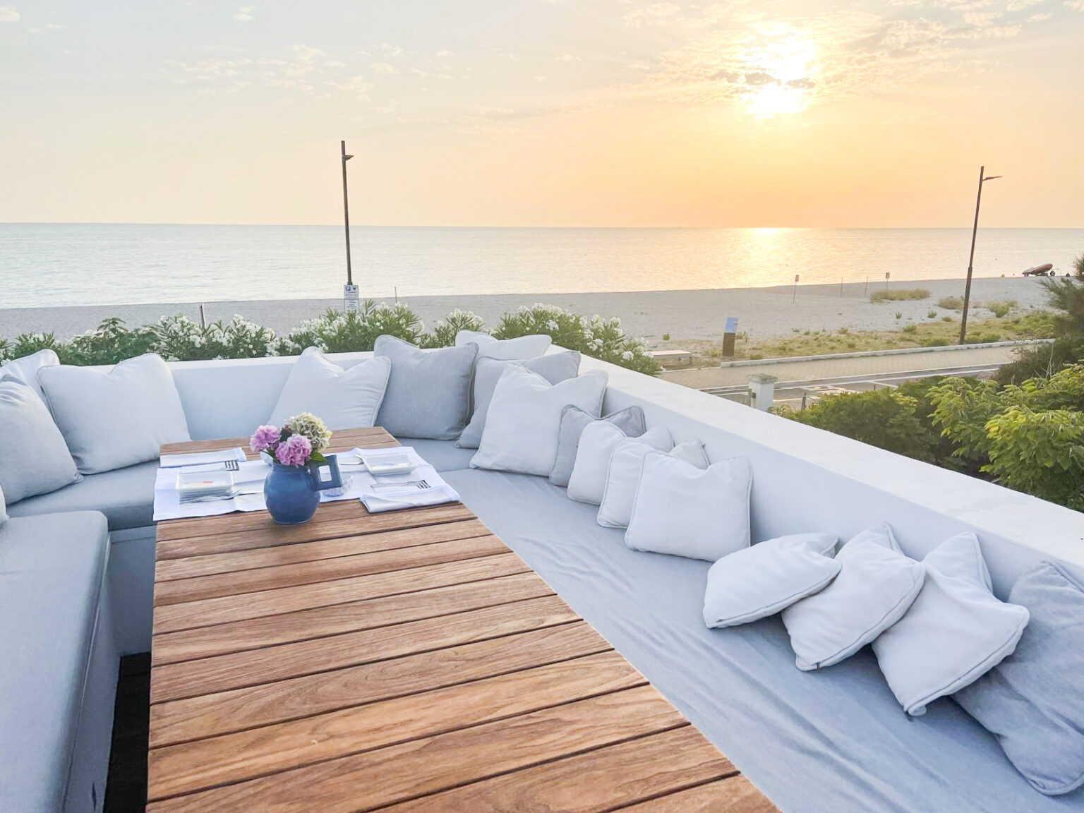 Rooftop seating area with a wooden table set for dining, surrounded by plush cushions, overlooking a serene beach at sunset. The scene captures a relaxing atmosphere with vibrant colors of the sky reflecting on the water. | Only on SopranoVillas