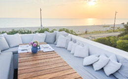 Rooftop seating area with a wooden table set for dining, surrounded by plush cushions, overlooking a serene beach at sunset. The scene captures a relaxing atmosphere with vibrant colors of the sky reflecting on the water.