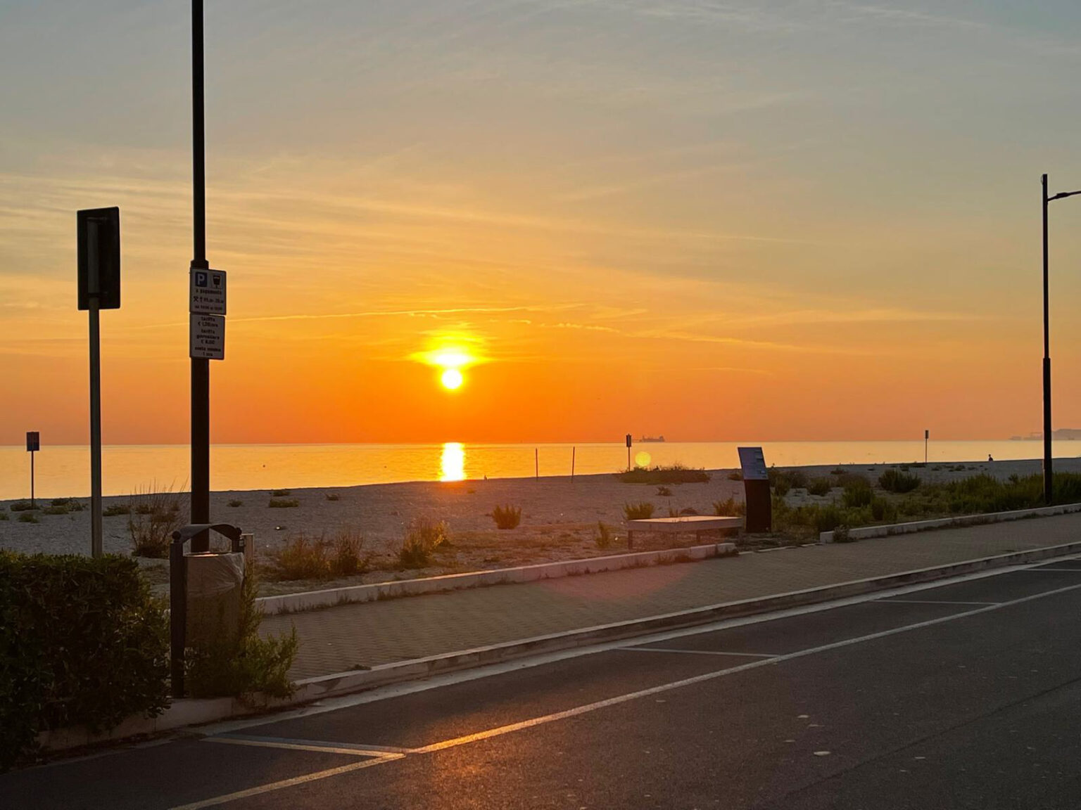 Sunset over a calm beach with golden hues reflecting on the water, surrounded by sandy shores and gentle vegetation, captured from a nearby roadway. | Only on SopranoVillas