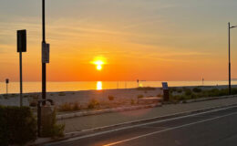 Sunset over a calm beach with golden hues reflecting on the water, surrounded by sandy shores and gentle vegetation, captured from a nearby roadway.