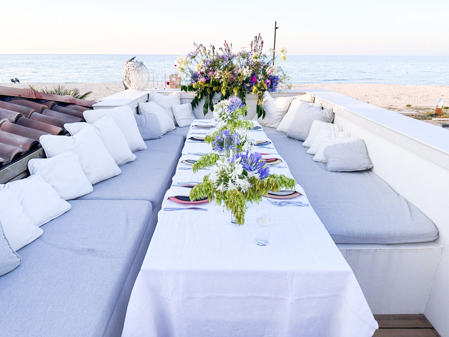 Elegant outdoor dining setup with a long table adorned with floral centerpieces, surrounded by comfortable cushions, overlooking a serene beach and sea. Perfect for gatherings and special occasions. | Only on SopranoVillas
