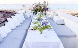 Elegant outdoor dining setup with a long table adorned with floral centerpieces, surrounded by comfortable cushions, overlooking a serene beach and sea. Perfect for gatherings and special occasions.