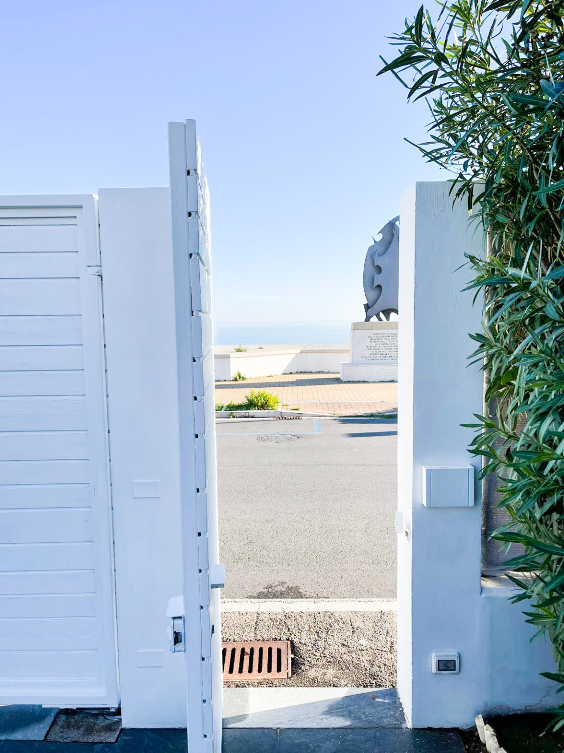 Open white gate leading to a scenic view of the ocean, framed by lush greenery, with a statue visible in the background and a clear blue sky above. | Only on SopranoVillas