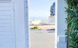 Open white gate leading to a scenic view of the ocean, framed by lush greenery, with a statue visible in the background and a clear blue sky above.