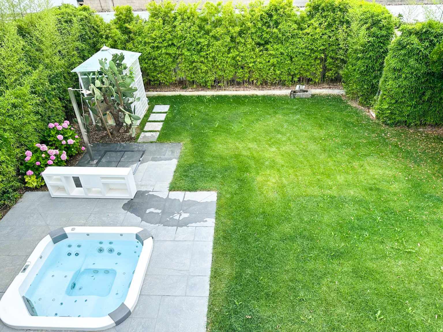 Aerial view of a landscaped backyard featuring a hot tub, a stone patio, a shower enclosure with a cactus, and vibrant pink flowers, surrounded by lush green hedges and a well-maintained lawn. | Only on SopranoVillas