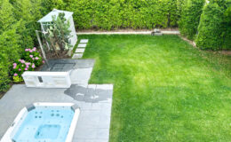 Aerial view of a landscaped backyard featuring a hot tub, a stone patio, a shower enclosure with a cactus, and vibrant pink flowers, surrounded by lush green hedges and a well-maintained lawn.