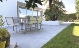 Modern outdoor patio featuring a sleek metal dining table with yellow chairs, surrounded by lush greenery and palm trees, ideal for alfresco dining and relaxation.