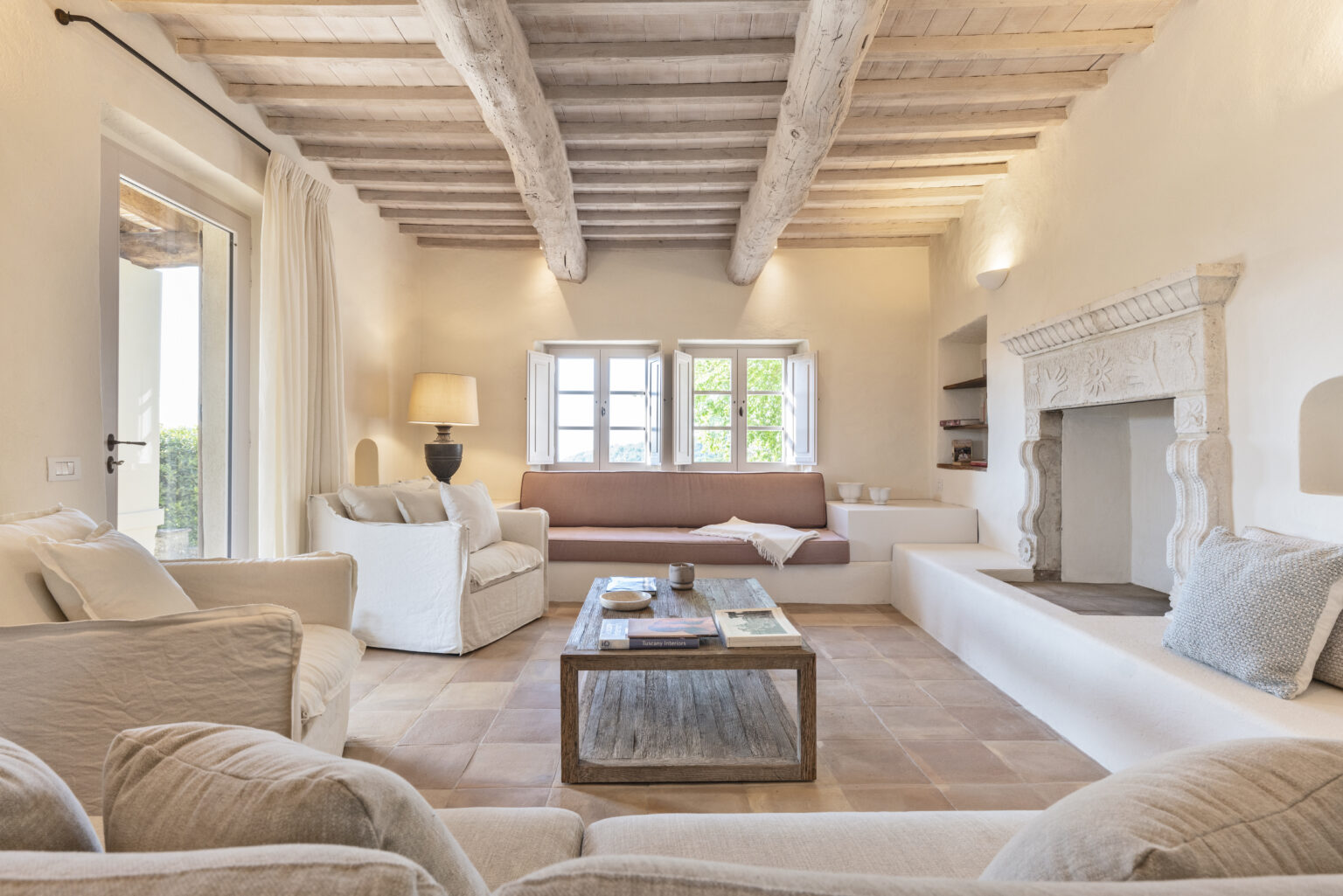 Spacious and inviting living room with rustic wooden beams, cozy white sofas, and a stone fireplace, featuring large windows that let in natural light and offer views of greenery. A wooden coffee table displays books and decorative items, enhancing the warm and welcoming atmosphere. | Only on SopranoVillas