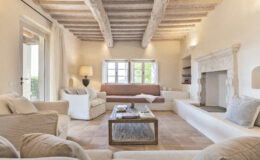 Spacious and inviting living room with rustic wooden beams, cozy white sofas, and a stone fireplace, featuring large windows that let in natural light and offer views of greenery. A wooden coffee table displays books and decorative items, enhancing the warm and welcoming atmosphere.