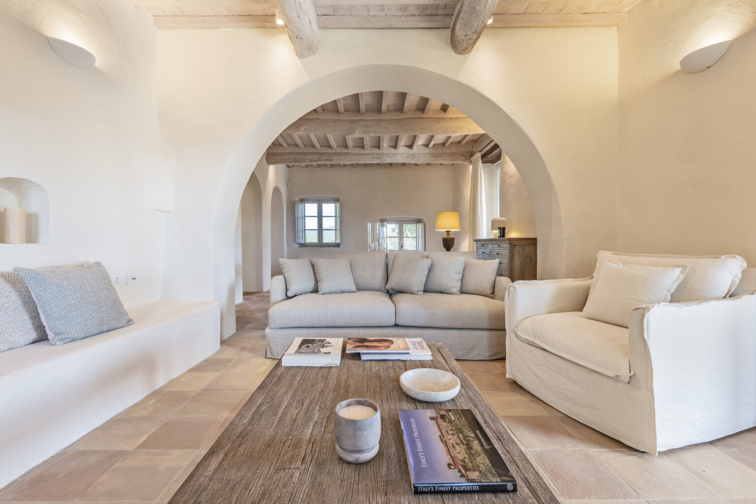 Cozy and modern living room featuring a light-colored sofa, comfortable armchair, and a rustic wooden coffee table. The space is enhanced by arched doorways, soft lighting, and natural textures, creating a warm and inviting atmosphere. Perfect for interior design inspiration. | Only on SopranoVillas