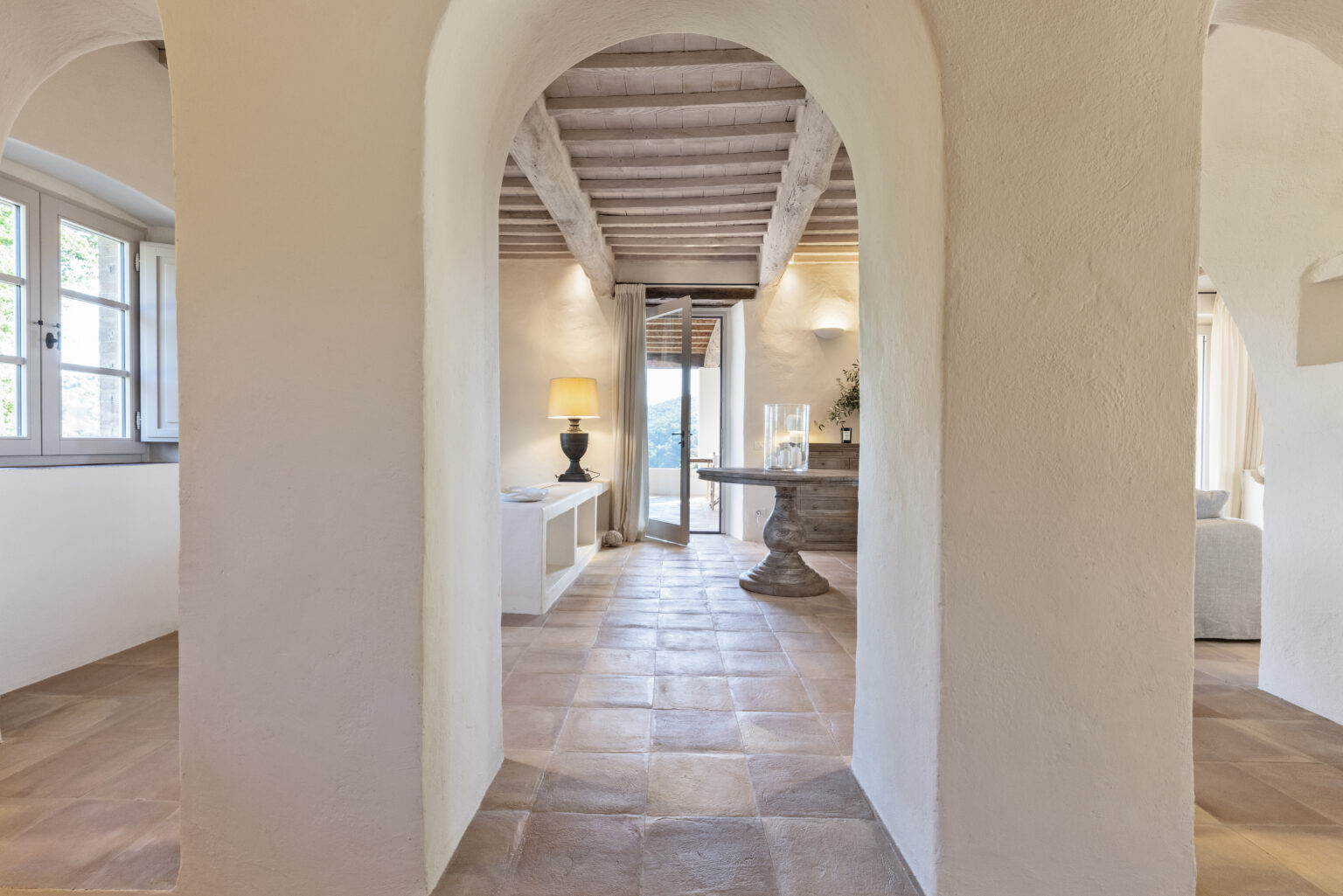 Interior view of a stylish, modern home featuring arched doorways, terracotta tiled flooring, and natural light streaming through large windows. A wooden table with decor and a lamp adds warmth to the inviting space. | Only on SopranoVillas