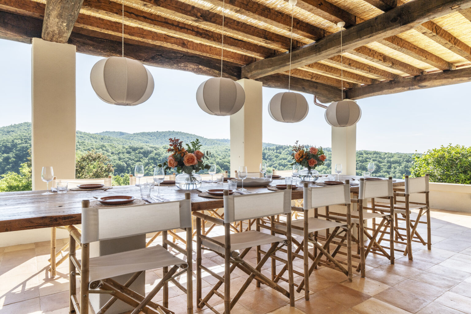 Outdoor dining area with a wooden table set for a meal, featuring elegant tableware and floral centerpieces. The space is adorned with hanging lanterns and offers a picturesque view of green hills under a clear blue sky. | Only on SopranoVillas