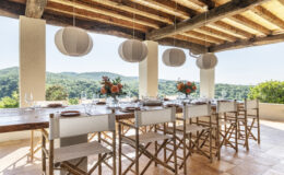 Outdoor dining area with a wooden table set for a meal, featuring elegant tableware and floral centerpieces. The space is adorned with hanging lanterns and offers a picturesque view of green hills under a clear blue sky.