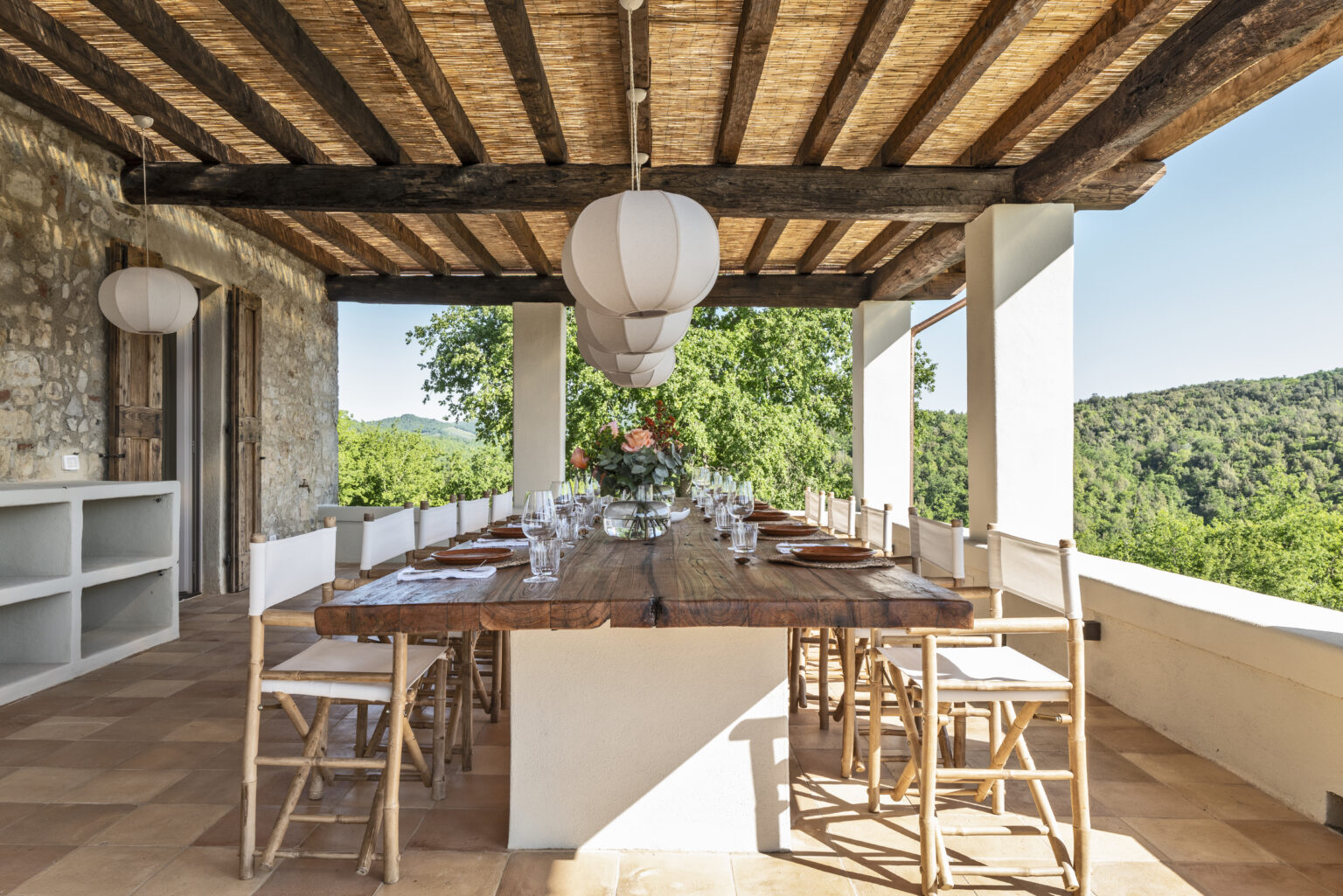 Outdoor dining area with a large wooden table set for a meal, surrounded by elegant chairs and a scenic view of lush greenery. The space features a thatched roof, wooden beams, and stylish pendant lighting, creating a warm and inviting atmosphere for gatherings. | Only on SopranoVillas