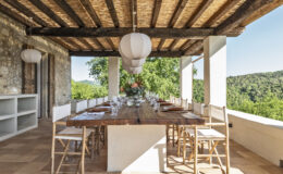 Outdoor dining area with a large wooden table set for a meal, surrounded by elegant chairs and a scenic view of lush greenery. The space features a thatched roof, wooden beams, and stylish pendant lighting, creating a warm and inviting atmosphere for gatherings.