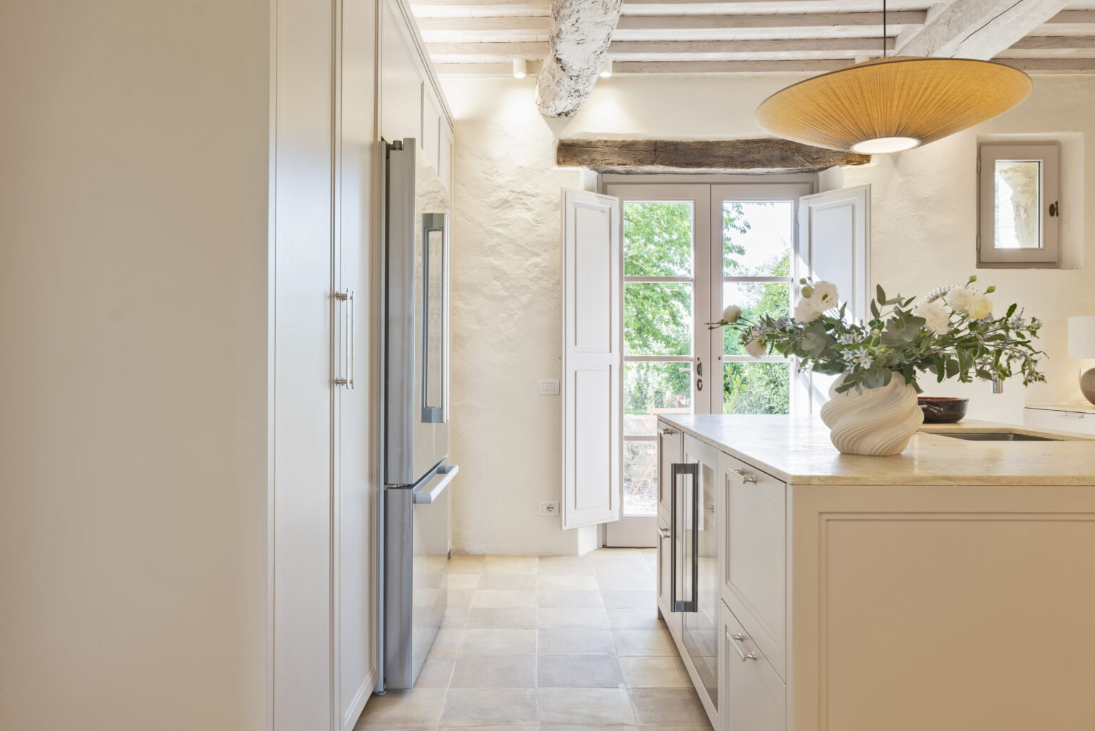 Modern kitchen interior featuring sleek cabinetry, stainless steel refrigerator, and a stylish pendant light. Natural light streams in through open French doors, illuminating a marble countertop adorned with a decorative vase of flowers. The space combines contemporary design with rustic elements, creating a warm and inviting atmosphere. | Only on SopranoVillas
