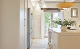 Modern kitchen interior featuring sleek cabinetry, stainless steel refrigerator, and a stylish pendant light. Natural light streams in through open French doors, illuminating a marble countertop adorned with a decorative vase of flowers. The space combines contemporary design with rustic elements, creating a warm and inviting atmosphere.