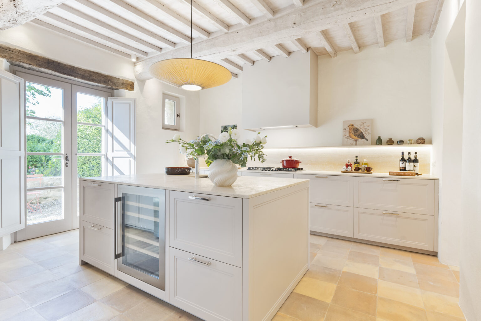 Modern kitchen interior featuring a spacious island with a wine cooler, elegant cabinetry, and a stylish pendant light. Natural light floods the room through large windows, highlighting the minimalist design and decorative elements, including fresh flowers and a red cooking pot on the stove. | Only on SopranoVillas