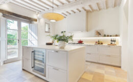 Modern kitchen interior featuring a spacious island with a wine cooler, elegant cabinetry, and a stylish pendant light. Natural light floods the room through large windows, highlighting the minimalist design and decorative elements, including fresh flowers and a red cooking pot on the stove.