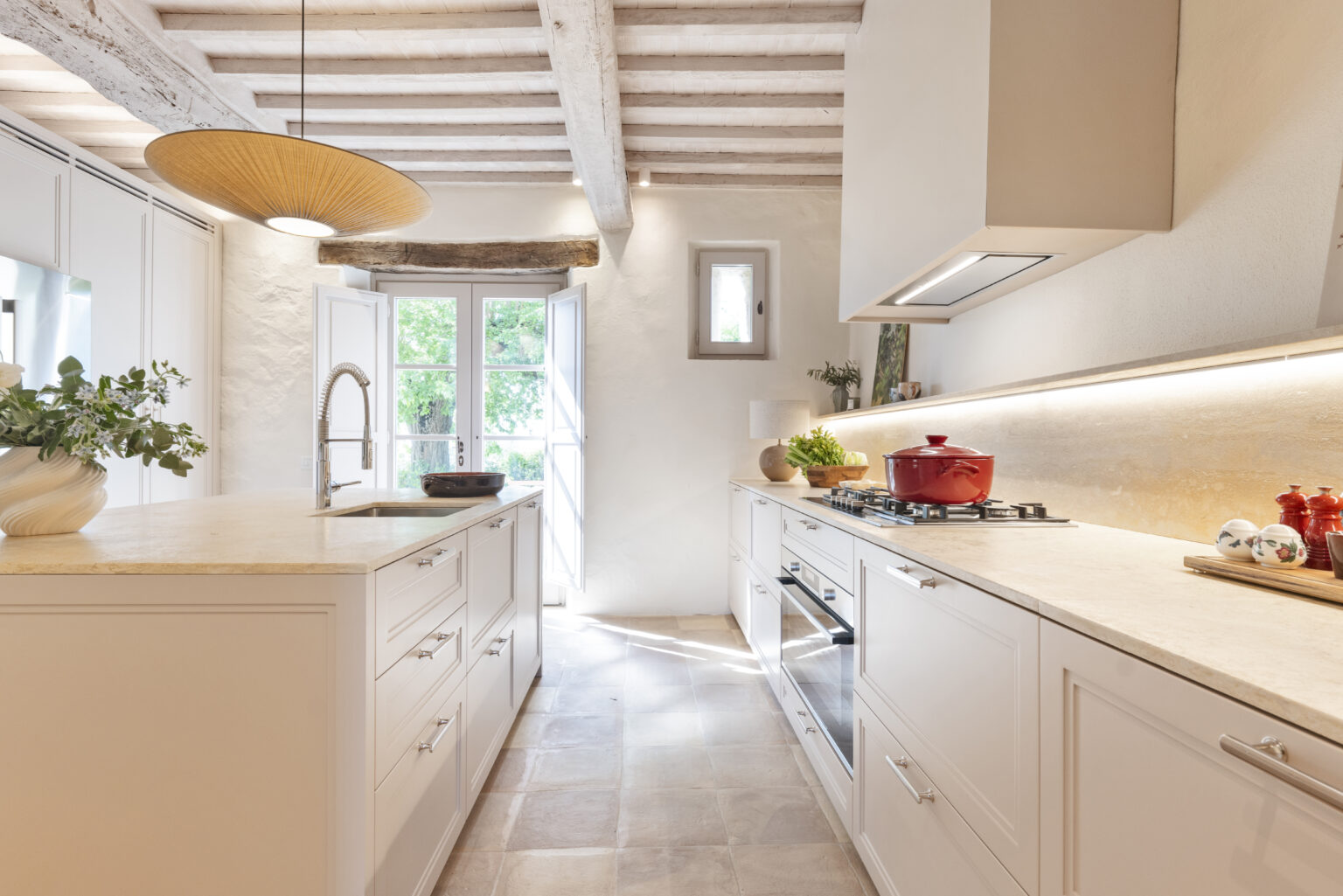 Modern kitchen interior featuring white cabinetry, a central island with a stone countertop, and stainless steel appliances. Natural light floods in through open French doors, highlighting a minimalist design with wooden beams and stylish decor elements. | Only on SopranoVillas