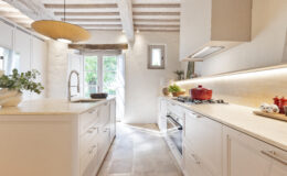 Modern kitchen interior featuring white cabinetry, a central island with a stone countertop, and stainless steel appliances. Natural light floods in through open French doors, highlighting a minimalist design with wooden beams and stylish decor elements.