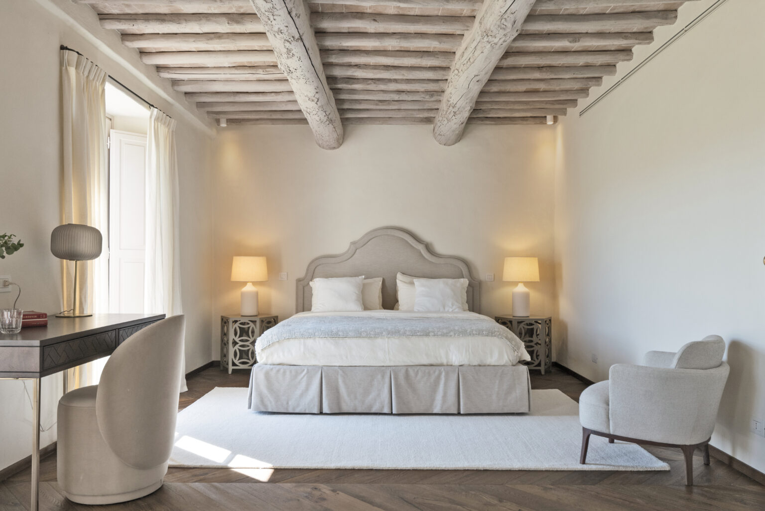 Modern bedroom interior featuring a plush bed with decorative pillows, soft lighting from two bedside lamps, a stylish desk with a unique chair, and large windows draped with light curtains. The room showcases wooden beams and a warm color palette, creating a cozy and inviting atmosphere. | Only on SopranoVillas