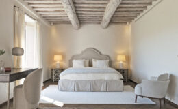 Modern bedroom interior featuring a plush bed with decorative pillows, soft lighting from two bedside lamps, a stylish desk with a unique chair, and large windows draped with light curtains. The room showcases wooden beams and a warm color palette, creating a cozy and inviting atmosphere.