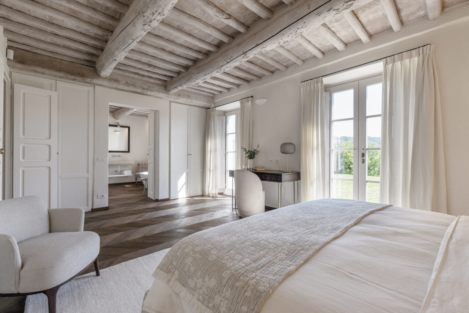 Bright and airy bedroom featuring wooden beams, large windows with sheer curtains, a cozy armchair, and a stylish desk, showcasing modern interior design and natural light. | Only on SopranoVillas