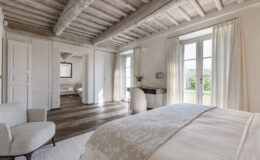 Bright and airy bedroom featuring wooden beams, large windows with sheer curtains, a cozy armchair, and a stylish desk, showcasing modern interior design and natural light.