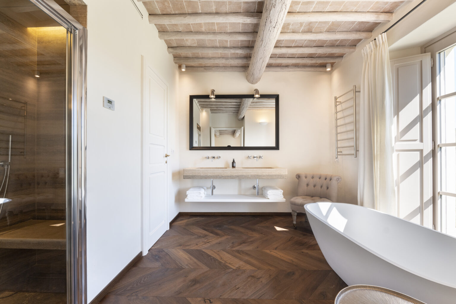 Modern bathroom featuring a freestanding bathtub, stylish vanity with a large mirror, and a walk-in shower. The space showcases elegant wooden flooring, exposed ceiling beams, and natural light streaming through the windows, creating a serene and luxurious atmosphere. | Only on SopranoVillas